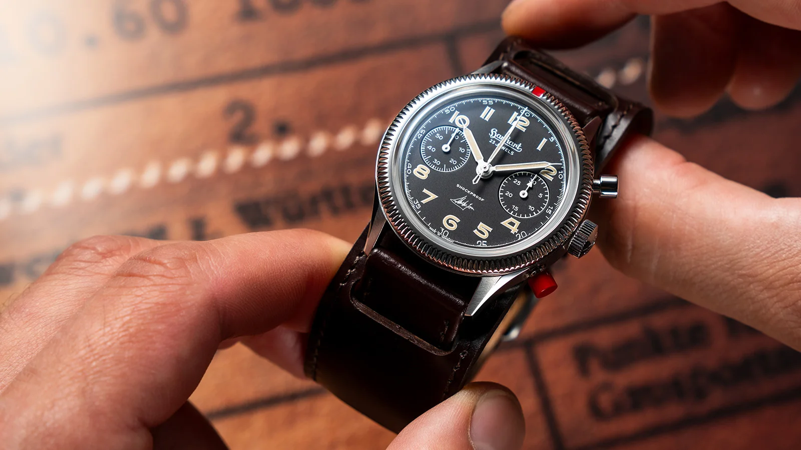Hanhart 417 ES Steve McQueen Limited Edition No. 1 zwischen zwei Händen: schwarzes Zifferblatt, rote Drückerkappe und braunes Unterlagenband.
